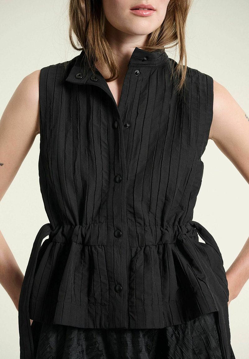 Femme portant un haut noir sans manches boutonné avec des coutures verticales et une taille cintrée nouée sur les côtés.