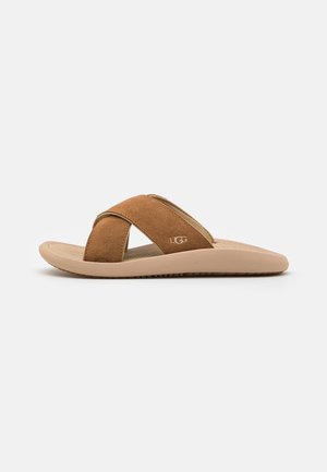 Brun ruskind slidesandal med krydsede stropper, beige sål og UGG-logo på siden, vist fra siden mod hvid baggrund.