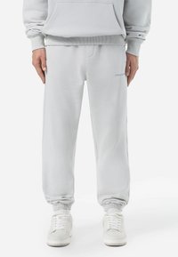 Pantalones de chándal de mezcla de algodón en gris claro con puños elásticos, cintura ajustable con cordón y un sutil detalle de logo en el lado izquierdo. Textura suave y corte relajado.