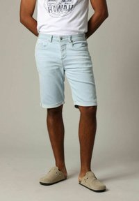 Shorts en denim bleu clair avec des ourlets retroussés, design à cinq poches, et fermeture à bouton en métal, associés à un T-shirt graphique blanc et des chaussures beiges.