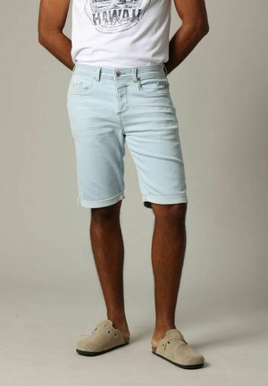 Shorts en denim bleu clair avec des ourlets retroussés, design à cinq poches, et fermeture à bouton en métal, associés à un T-shirt graphique blanc et des chaussures beiges.