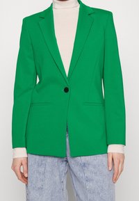 Blazer verde realizzato in tessuto liscio con rever a punta, chiusura con un bottone e due tasche frontali. Indossato sopra un dolcevita beige.