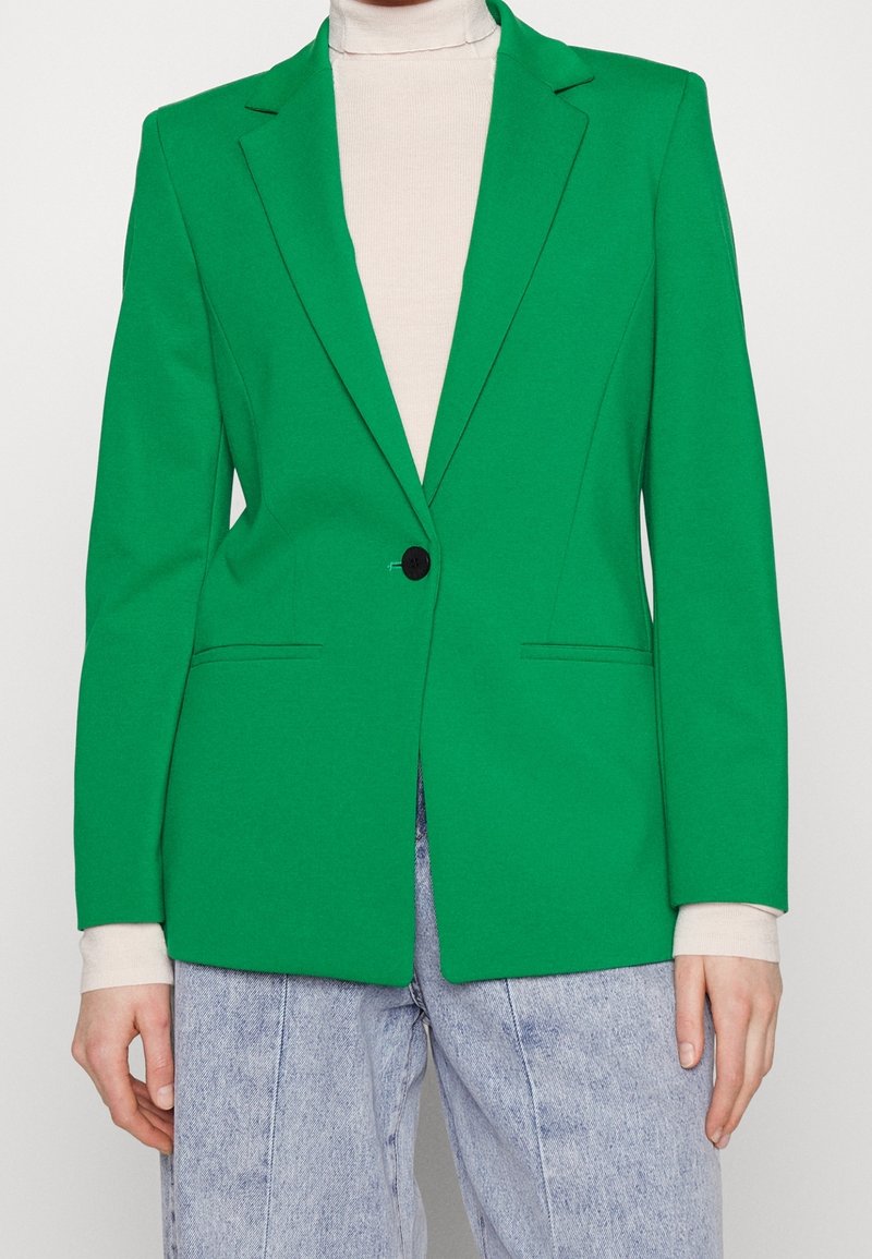 Blazer verde realizzato in tessuto liscio con rever a punta, chiusura con un bottone e due tasche frontali. Indossato sopra un dolcevita beige.