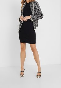 Blazer a quadri bianco e nero con piping bianco a contrasto, indossato sopra un vestito nero a coste. Tacchi alti a strisce bianche e nere completano il look.