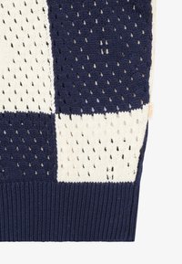 Gestrickter Pullover mit kariertem Muster in Marineblau und Creme, strukturierter offen gewebter Oberfläche und einem gerippten Marineblauen Saum. Seitenetikett sichtbar.