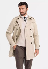 Beżowy trencz z podwójnym zapięciem, pagonami na ramionach i eleganckim krojem. Nosi się go na lekkim swetrze i szarych spodniach.