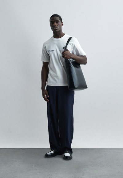 Άνδρας που στέκεται μπροστά σε έναν απλό τοίχο φορώντας λευκό t-shirt με γραφικά, μπλε παντελόνι με ρίγες και φαρδιά μπατζάκια, μαύρα και λευκά loafers, και κρατώντας μια μαύρη δερμάτινη τσάντα tote.