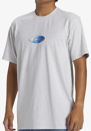 Homme portant un t-shirt gris clair à manches courtes avec un logo ovale bleu et blanc sur la poitrine, associé à un jean bleu.