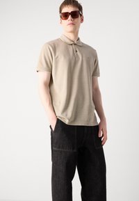 Beige polo-skjorte med to-knappers knappestolpe, korte ermer og en subtil tekstur. Kombinert med mørke jeansbukser med sidelommer.