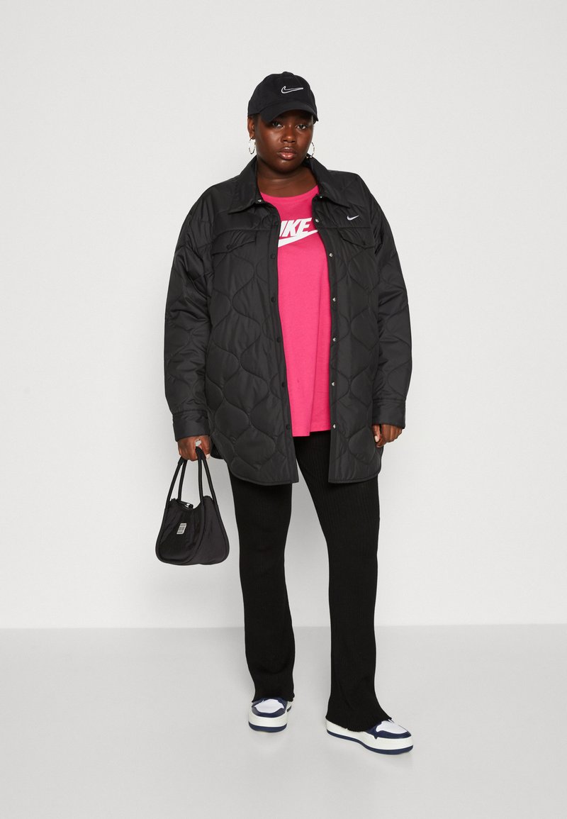 Veste matelassée noire sur un t-shirt rose Nike, associée à un pantalon noir côtelé et des baskets blanches. Accessoire comprenant un petit sac noir.