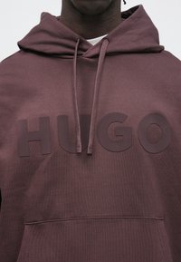 Primo piano di una persona che indossa una felpa con cappuccio marrone con una tasca frontale e il logo "HUGO" stampato sul petto.