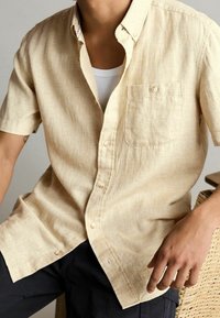 Camisa de botones de manga corta color beige con un patrón texturizado, un bolsillo en el pecho y una tela suave. Camiseta blanca visible debajo.