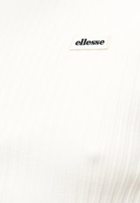 Κοντινή λήψη λευκού υφάσματος με ραβδώσεις και μικρό μαύρο έμβλημα λογότυπου "ellesse" στο πάνω αριστερό μέρος.