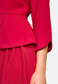 Fuchsia Kleid mit Peplum-Design, langen Ärmeln und weichem Stoff. Falten sind an der Taille sichtbar und zeigen eine glatte Textur.