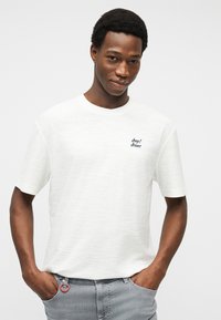 Νέος άντρας με κοντά σγουρά μαλλιά, που φοράει λευκό T-shirt "Joop! Jeans" και γκρι παντελόνι, στέκεται με τα χέρια στις τσέπες.