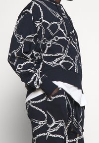 Marinblå hoodie med vit kedjemönster, har en dragsnodd huva, ribbstickade ärmslut och en avslappnad passform. Kombinerad med matchande byxor.