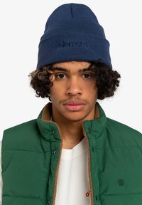 Beanie a coste color blu navy con logo "element", indossato con un gilet imbottito verde con colletto in pile marrone. Lo sfondo è bianco semplice.