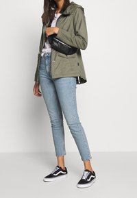 Person med grön hooded jacka, vit t-shirt, ljusblå jeans, svarta och vita sneakers, håller en svart axelväska i midjehöjd.