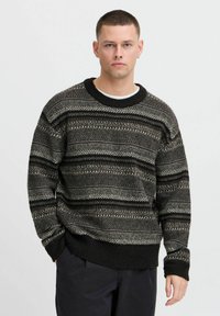 Black and gray patterned sweater with a round neckline, featuring a mix of horizontal stripes and textures, worn over a white shirt.