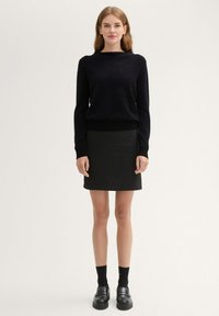 Schwarzer strukturierten Pullover mit weitem Ausschnitt, kombiniert mit einem schwarzen Bleistiftrock. Das Model trägt schwarze Socken und Loafers. Schlichtes, monochromatisches Design.