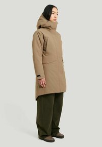 Beige vattentät parka med hög krage och huva, med två framfickor och svarta accenter, kombinerad med olivgröna vida byxor.
