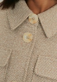 Bruin geweven jas met een gestructureerde afwerking, voorzien van een kraag en twee crèmekleurige knopen aan de voorkant. Twee zakken zichtbaar.