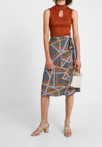 Top sin mangas marrón de canalé con un corte en forma de llave, combinado con una falda midi estampada que presenta diseños geométricos en azul, naranja y negro. Bolso transparente.