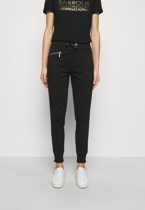 Modèle portant un t-shirt noir Barbour International et un jogger noir avec poignets et poche zippée dorée, associé à des baskets blanches.