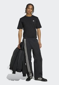 Čierne tričko s bielym logom, čierne cargo nohavice s tromi bielymi pruhmi, drží čiernu prešívanú bundu, obuté čierne tenisky.