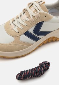 Beżowe i jasnoszare sneakersy z zamszowymi akcentami, niebieskim logo i teksturowaną gumową podeszwą. W komplecie sprężynowy czarny i czerwony sznurowadło.