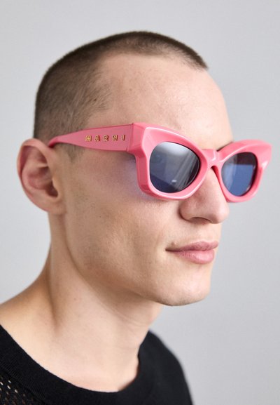 Lyserøde cat-eye solbriller med vinklet stel, blåtonede linser og guldbranding på siden. Mat plastfinish, dristigt og geometrisk design.
