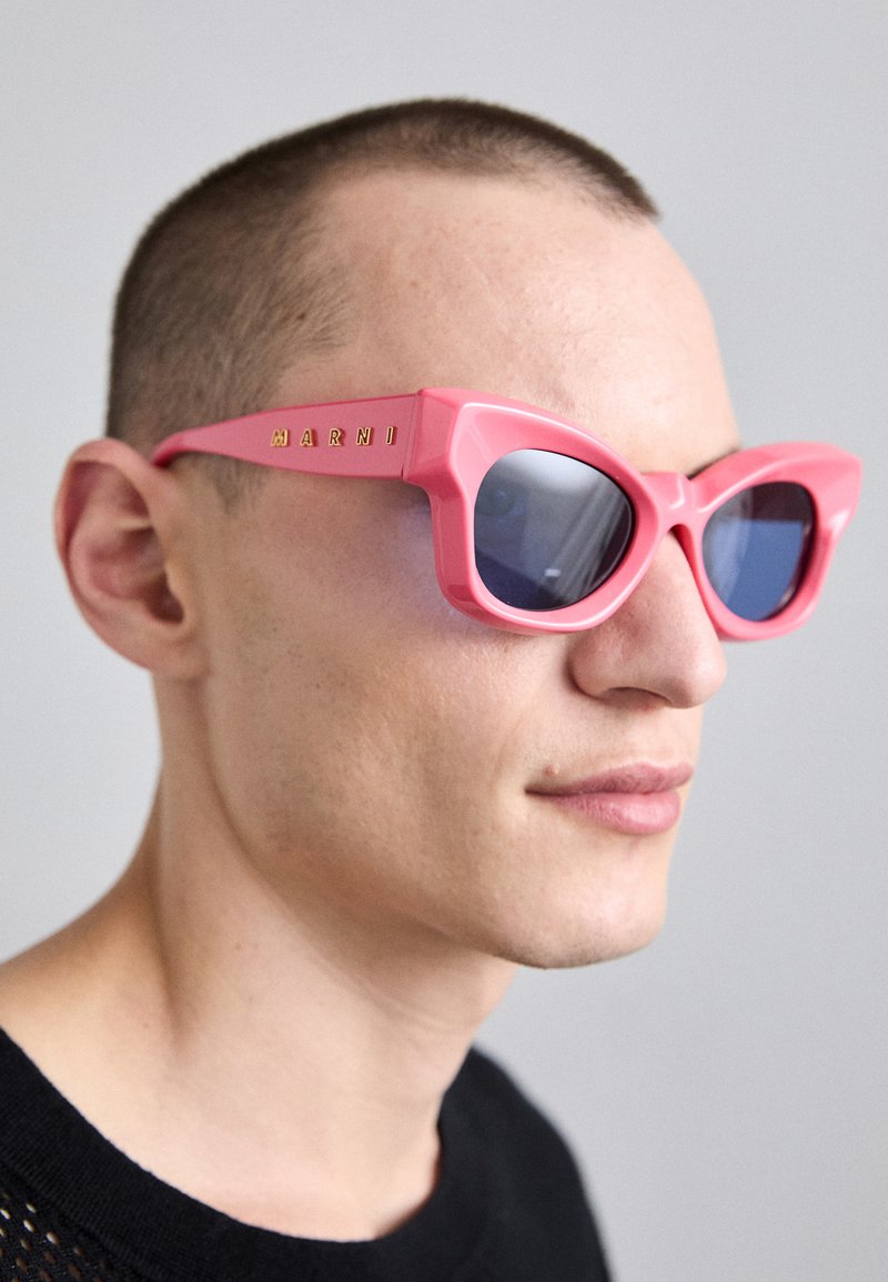 Occhiali da sole a forma di occhio di gatto rosa con montatura angolare, lenti di colore blu e marchio dorato sul lato. Finitura in plastica opaca, design audace e geometrico.