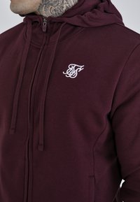 Bordeaux kleurige hoodie met rits in zachte stof beschikt over een wit logo op de borst, verstelbare trekkoorden en een ontspannen pasvorm met een kangoeroezak.