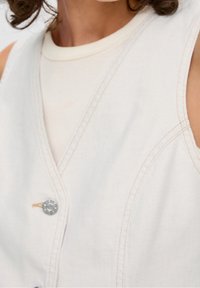 Weißes Jeanswesten mit V-Ausschnitt, versehen mit kontrastierenden orangen Nähten und runden silbernen Knöpfen. Über einem hellen Tanktop getragen.