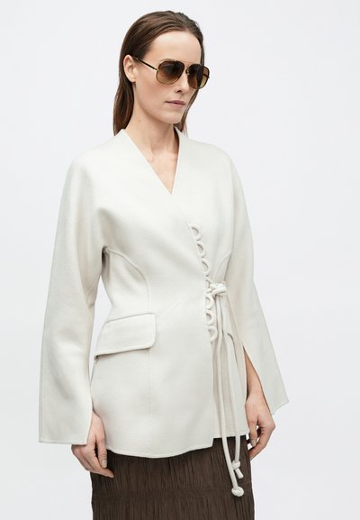 Λευκό σακάκι-περιτύλιγμα με μακριά μανίκια, πλαϊνή τσέπη και διακοσμητικούς δεσμούς. Το μοντέλο φοράει καφέ φούστα και μεγάλα γυαλιά ηλίου με καφέ απόχρωση.