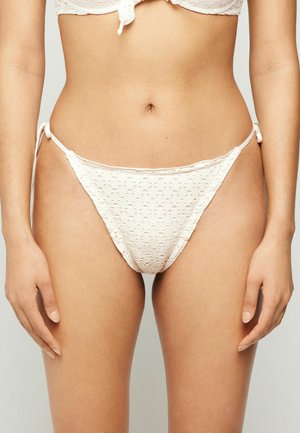 Femme portant un bas de bikini blanc en tissu ajouré avec des liens sur les côtés et un haut de bikini assorti avec un nœud à l'avant.