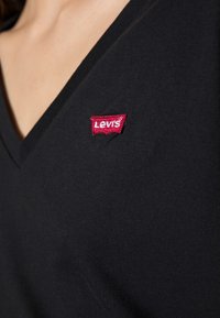 Černé V-krční tričko s texturovanou tkaninou, na výstřihu má červenou vyšitou nášivku loga Levi's.