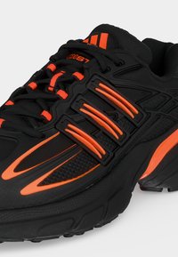 Baskets de sport noires avec des bandes orange vif, des accents le long de la semelle et des lacets noirs passés dans des œillets.