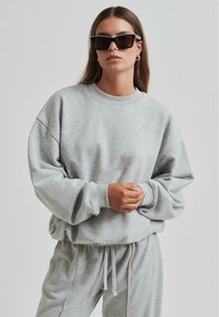 Sudadera gris con un ajuste relajado, cuello acanalado y mangas largas. Combinada con pantalones de chándal grises y gafas de sol negras. Textura suave.