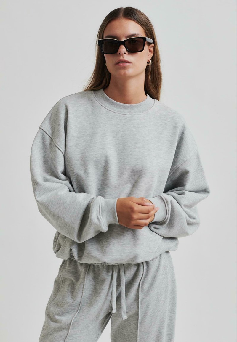 Sudadera gris con un ajuste relajado, cuello acanalado y mangas largas. Combinada con pantalones de chándal grises y gafas de sol negras. Textura suave.