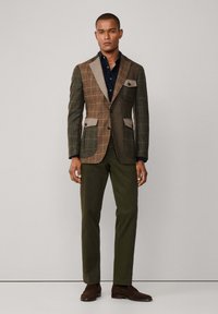Bruin geruit blazer met contrasterende lichtgrijze revers, twee voorzakken, gecombineerd met donkergroene broek en bruine schoenen.