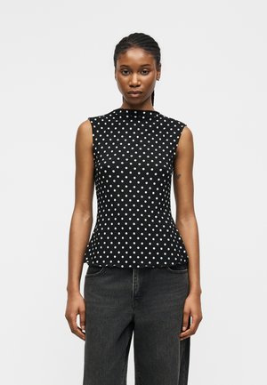 Kvinde iført sort ærmeløs top med hvide polkaprikker og højttaljede mørkegrå jeans, stående mod en ensfarvet hvid baggrund.