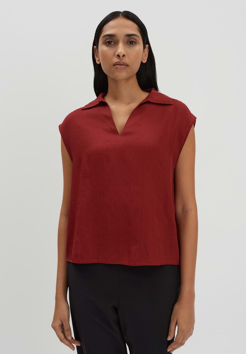 Blusa rossa a maniche corte con scollo a V e colletto. Tessuto liscio, vestibilità morbida e orlo dritto. Abbinata a pantaloni neri per un contrasto.