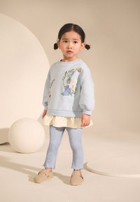 Jasnoniebieska bluza z haftem kwiatowym, noszona na kremowej spódnicy, w zestawie z niebieskimi legginsami i jasnobrązowymi wsuwanymi butami.