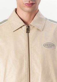 Beige Lederjacke mit Reißverschluss, spitzem Kragen und strukturierten Oberflächen. Enthält ein graues, gesticktes Logo auf der Brust.