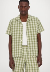 Κοντομάνικο πουκάμισο σε πράσινη και λευκή καρώ με καμπυλωτό γιακά, δύο μπροστινές τσέπες και άνετη εφαρμογή. Φοράει πάνω από ένα λευκό αμάνικο.