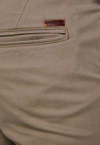 Pantalons en coton beige avec une poche latérale et une étiquette de logo cousue. Texture lisse avec des plis subtils et des éléments de design ajustés.