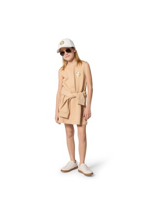 Robe sans manches beige avec une taille nouée, logo doré, casquette blanche et lunettes de soleil. Les chaussures sont blanches avec un design texturé et des semelles marron.