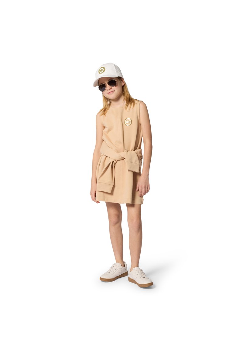 Μπεζ αμάνικο φόρεμα με ζώνη δεμένη στη μέση, χρυσό λογότυπο, άσπρο καπέλο και γυαλιά ηλίου. Τα παπούτσια είναι άσπρα με υφασμάτινο σχέδιο και καφέ σόλες.