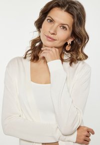 Krém színű kardigán nyitott elülső résszel és hosszú ujjakkal, fehér póló fölött viselve. Nagy, textúrált rózsaszín fülbevalókkal kísérve. Puha, könnyű anyag.